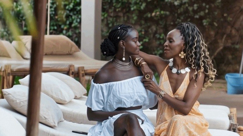 Two women sitting close together on an outdoor daybed, wearing handmade jewelry